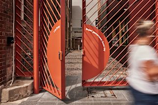 Red metal gate with the phrases "Have a good day" and "Bujari gamarruwa" partially open. Blurred person walking past on a paved path.