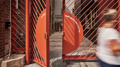 Red metal gate with the phrases "Have a good day" and "Bujari gamarruwa" partially open. Blurred person walking past on a paved path.
