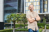 A man with short hair and a beard stands outside with arms crossed, smiling in front of a modern building with glass windows and trees.