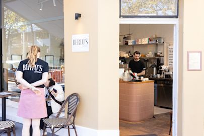 Two women sit outside a café called Berto’s Espresso, while a barista prepares drinks behind the counter inside.