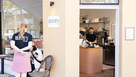 Two women sit outside a café called Berto’s Espresso, while a barista prepares drinks behind the counter inside.