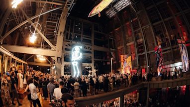 A large indoor venue with a crowd of people observing illuminated modern art installations and large colorful banners hanging on industrial walls.