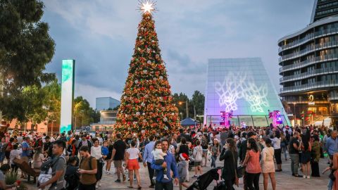 A large crowd gathers around a tall, decorated Christmas tree in a public square at dusk, with modern buildings in the background.