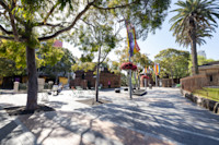 A sunlit plaza with empty tables and chairs, trees providing shade, colorful flags hanging, and a few people in the background.