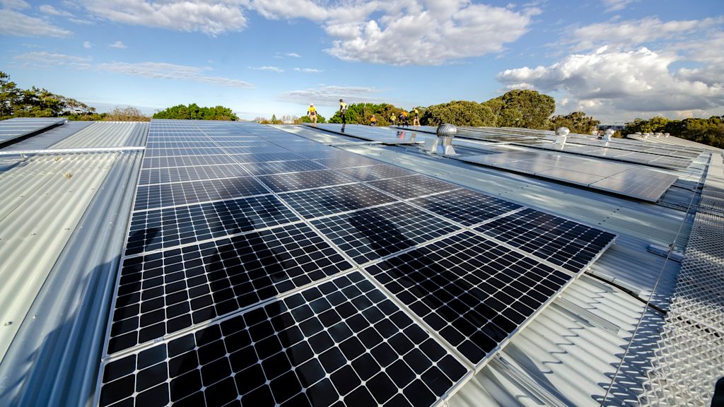 A large array of solar panels is installed on a metal rooftop under a partly cloudy sky, surrounded by trees and greenery.