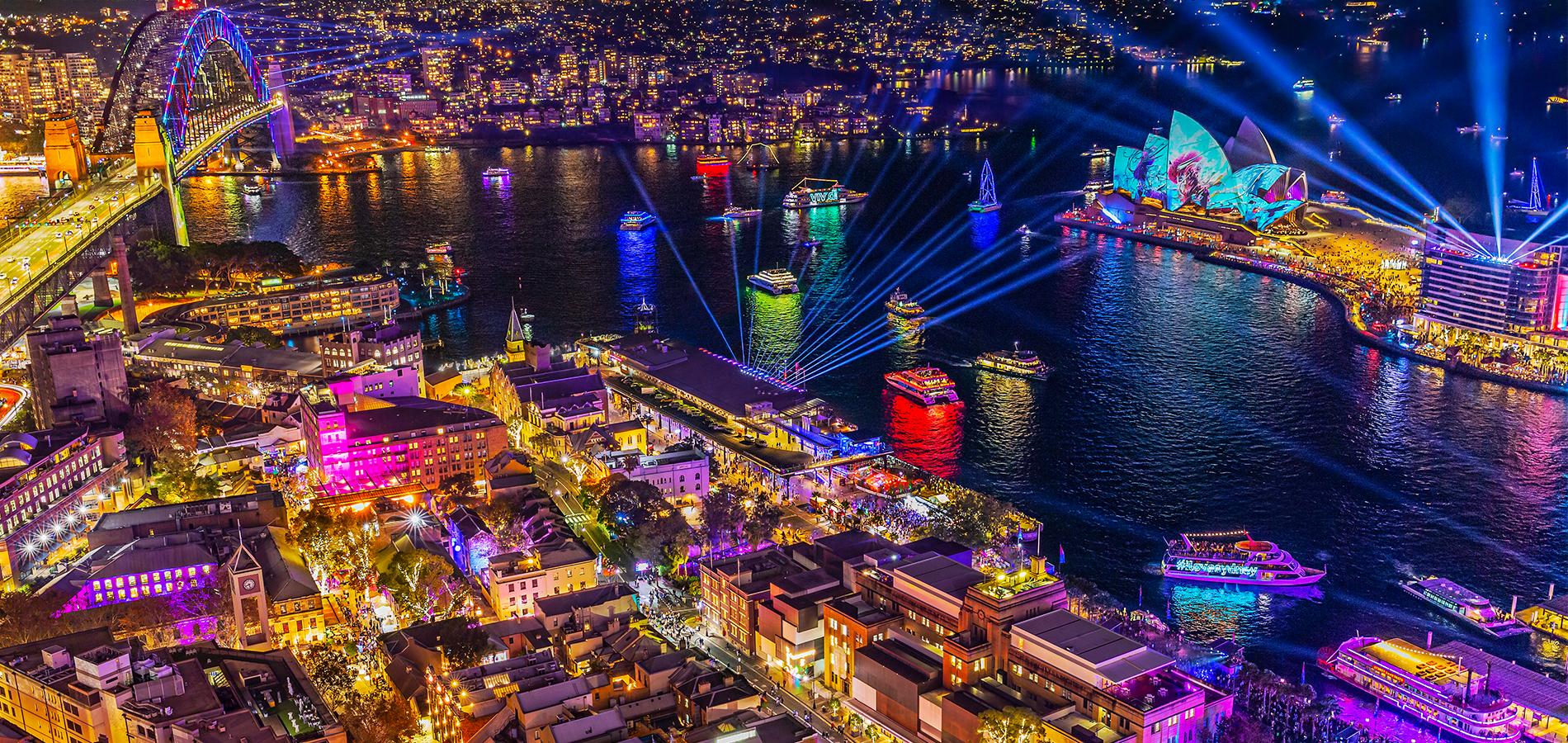 Aerial view of Sydney Harbour during Vivid at night. The Sydney Opera House, Harbour Bridge, and buildings are illuminated with light displays and projection art, while crowds gather around Circular Quay and The Rocks. Image courtesy of Vivid.