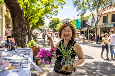 A woman smiles while holding a pot with pink flowers and another with a green plant at an outdoor market. Tables with various items for sale line the street, with people and trees in the background.