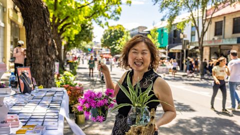 A woman smiles while holding a pot with pink flowers and another with a green plant at an outdoor market. Tables with various items for sale line the street, with people and trees in the background.