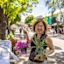 A woman smiles while holding a pot with pink flowers and another with a green plant at an outdoor market. Tables with various items for sale line the street, with people and trees in the background.