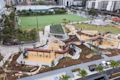 Aerial view of a modern playground with slides, climbing structures, swings, and nearby sports fields, surrounded by urban buildings and landscaping.