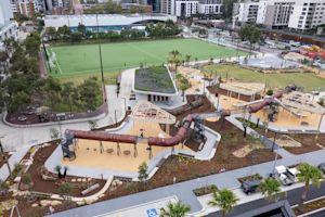 Aerial view of a modern playground with slides, climbing structures, swings, and nearby sports fields, surrounded by urban buildings and landscaping.