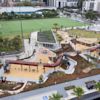 Aerial view of a modern playground with slides, climbing structures, swings, and nearby sports fields, surrounded by urban buildings and landscaping.