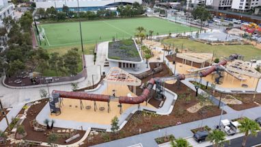 Aerial view of a modern playground with slides, climbing structures, swings, and nearby sports fields, surrounded by urban buildings and landscaping.