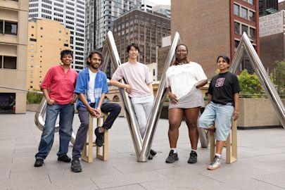 Five people pose on and beside a metal sculpture in an urban plaza, with tall buildings in the background.