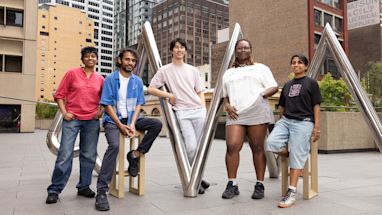 Five people pose on and beside a metal sculpture in an urban plaza, with tall buildings in the background.