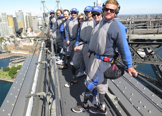 Jade 'Red' Wheatley, pro-adaptive surfer with group of amputee surfers at the summit of the Sydney Harbour Bridge Climb. They wear sunglasses and a safety harness, with city skyline in the background. Image courtesy of Sydney Bridge Climb.