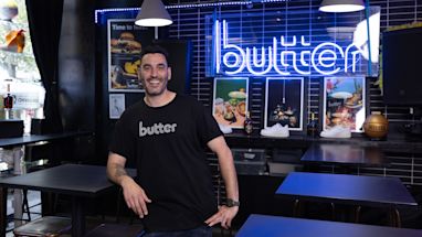 A man wearing a black t-shirt branded "Butter" stands and smiles inside a modern restaurant with a neon "butter" sign and food posters on the wall behind him.