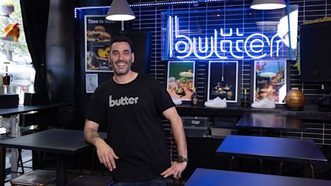 A man wearing a black t-shirt branded "Butter" stands and smiles inside a modern restaurant with a neon "butter" sign and food posters on the wall behind him.