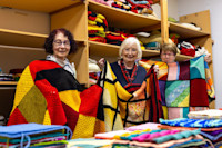 Three older women stand in front of shelves holding colourful knitted blankets, each displaying a handmade patchwork wrap in a room filled with folded knitted items.