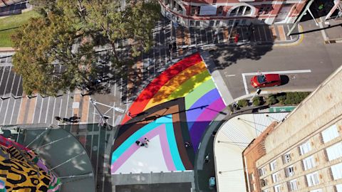 Aerial view of the Bourke and Campbell Street intersection in Darlinghurst painted with a Progress Pride flag, with pedestrians walking nearby, a red car parked on the side, and buildings and trees in the surroundings.