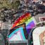 Aerial view of the Bourke and Campbell Street intersection in Darlinghurst painted with a Progress Pride flag, with pedestrians walking nearby, a red car parked on the side, and buildings and trees in the surroundings.