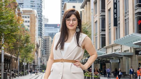 A woman wearing a cream-colored vest and pants stands on a city street lined with tall buildings and trees, smiling at the camera.