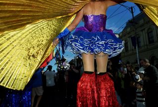 A performer in colorful costume with golden wings stands elevated in front of a crowd during an outdoor event.