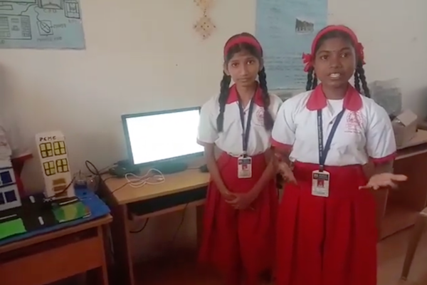 Two girls in school uniform explain their computing project next to a laptop and paper model of the project.