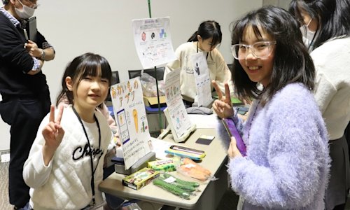 Two girls display posters about their computing project at a busy event.