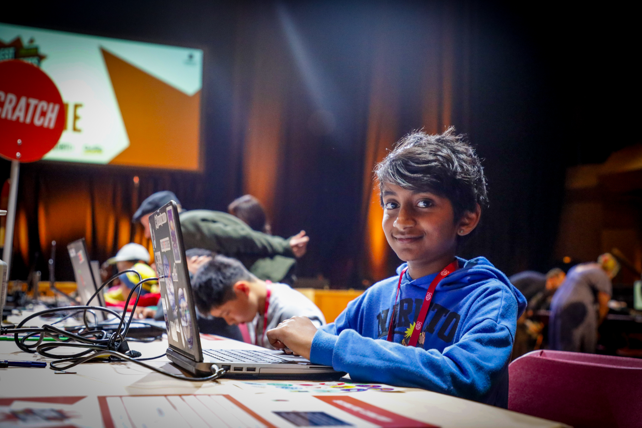 A young boy sits at a desk with a laptop, and smiles at the camera.
