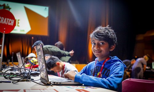 A young boy sits at a desk with a laptop, and smiles at the camera.