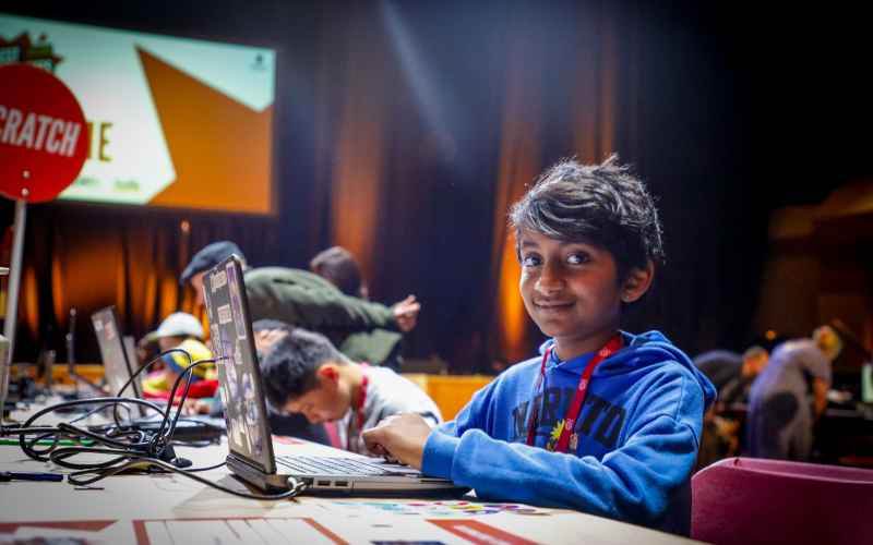 A young boy sits at a desk with a laptop, and smiles at the camera.