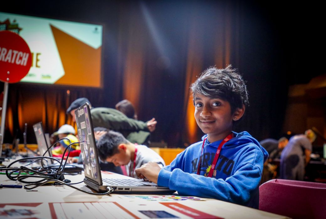 A young boy sits at a desk with a laptop, and smiles at the camera.