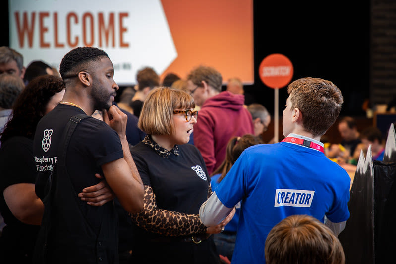 A young person in a blue t-shirt shows their technology project to two judges in black t-shirts at a Coolest Projects in-person event.