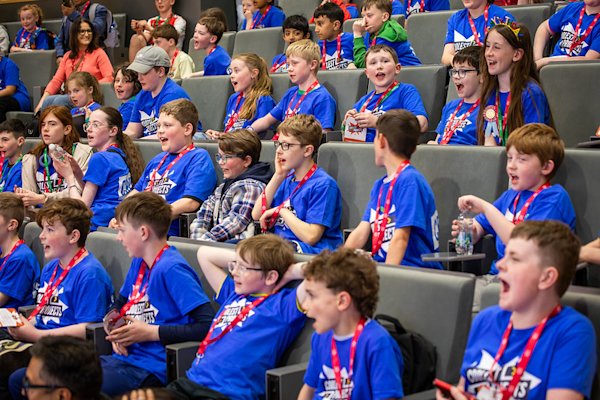 A group of young people in blue t-shirts and red lanyards cheer in an auditorium.