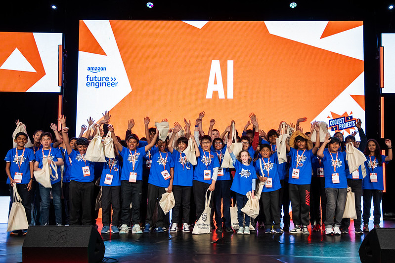 A group of young people in blue t-shirts cheer on stage.