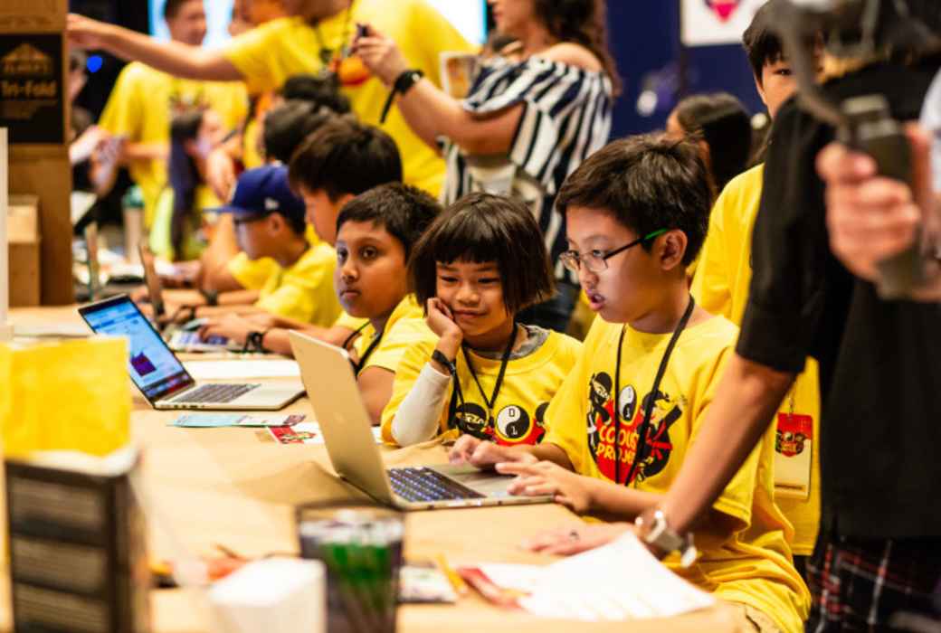 A group of girls and boys work on a project at a laptop at a table.