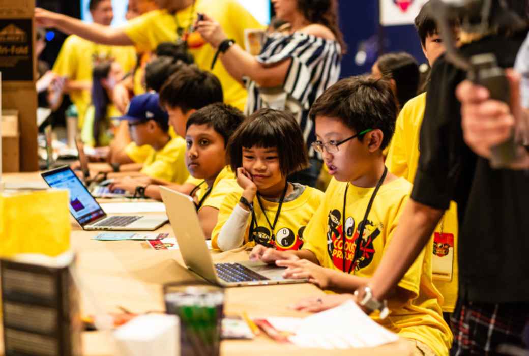 A group of girls and boys work on a project at a laptop at a table.