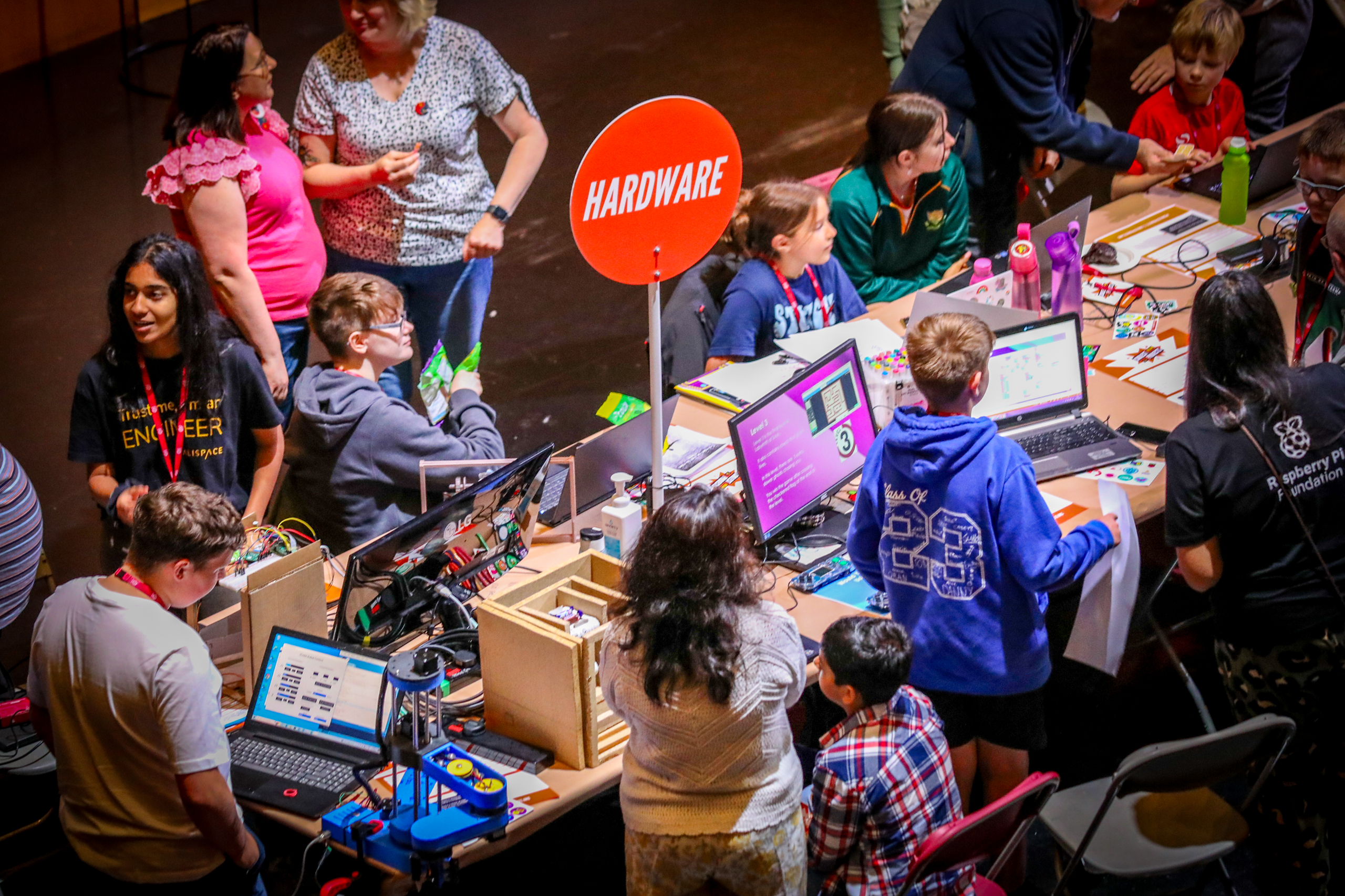 Young people sit at a table with laptops and computers at a busy event.