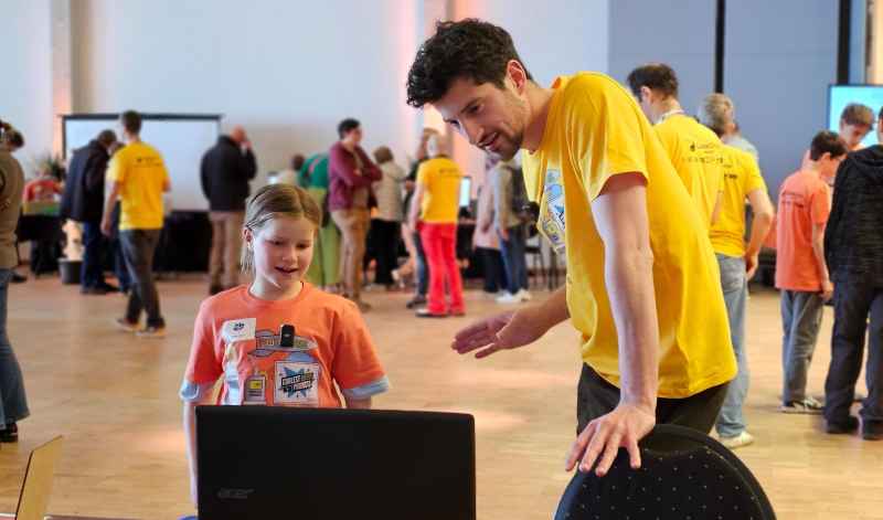 A young person and an adult mentor look at a laptop at a busy event.