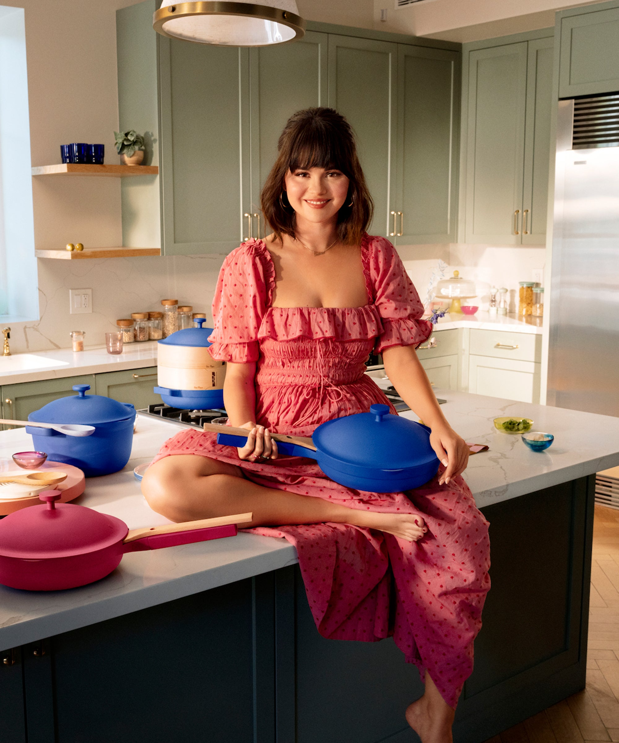 Selena Gomez in a pink dress, sitting on a kitchen counter with a blue frying pan in her lap.