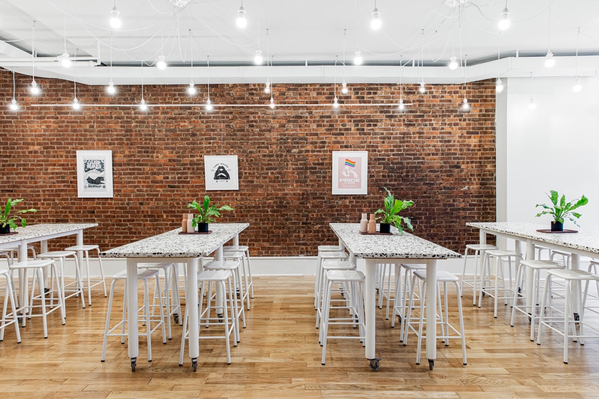 Maven Clinic office renovation interior design by Basicspace. All hands assembly white seating enveloped in the texture of brick and playful terrazzo custom tables on wheels. Hanging pendant lights, green ferns, and minimal art track finish the space