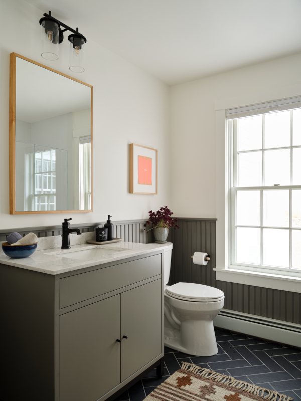 This beautiful bathroom features a flush paneled vanity with a warm grey colored paint, matte black hardware and white quartz countertop. The deep coloring continues on wainscoting that wraps the entirety of the room with warm white walls above. Herringbone tile covers the floor and makes the natural wood frame of the mirror pop. 