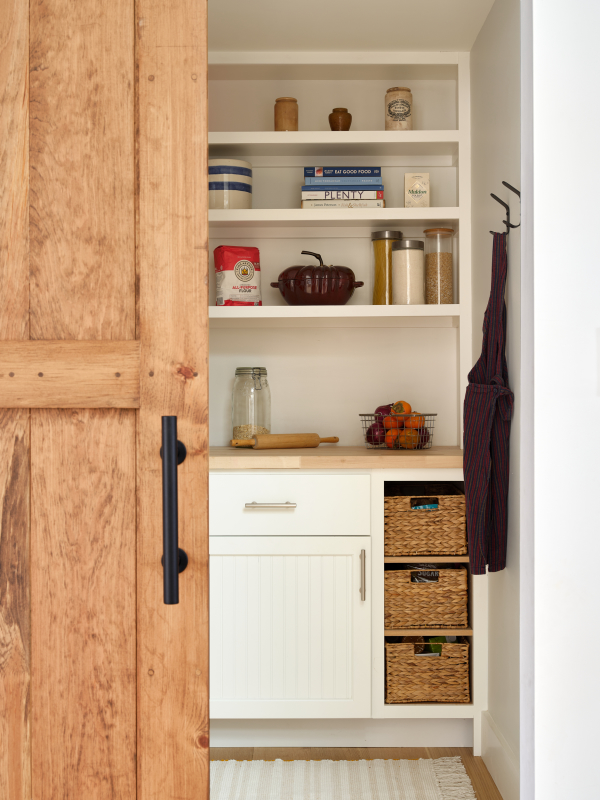 This walk in pantry features a sliding barn door, which reveals the elegant storage system. Open shelving sits above a wooden butcher block countertop and paneled cabinet storage below.