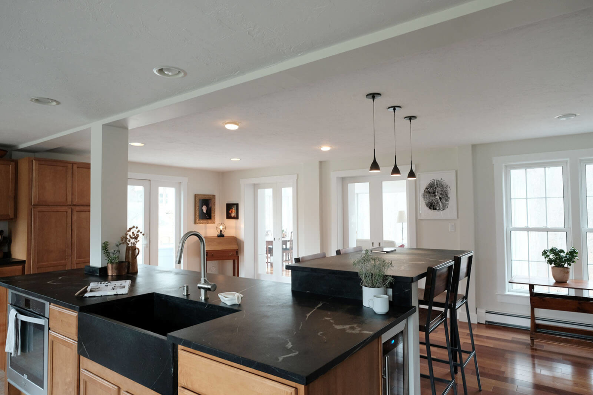 Jerusalem Road House residential interior design by Basicspace. Displaying the relationship between the kitchen and partial dining space. Open kitchen with bar height seating, farmhouse sink, and black soapstone counters.