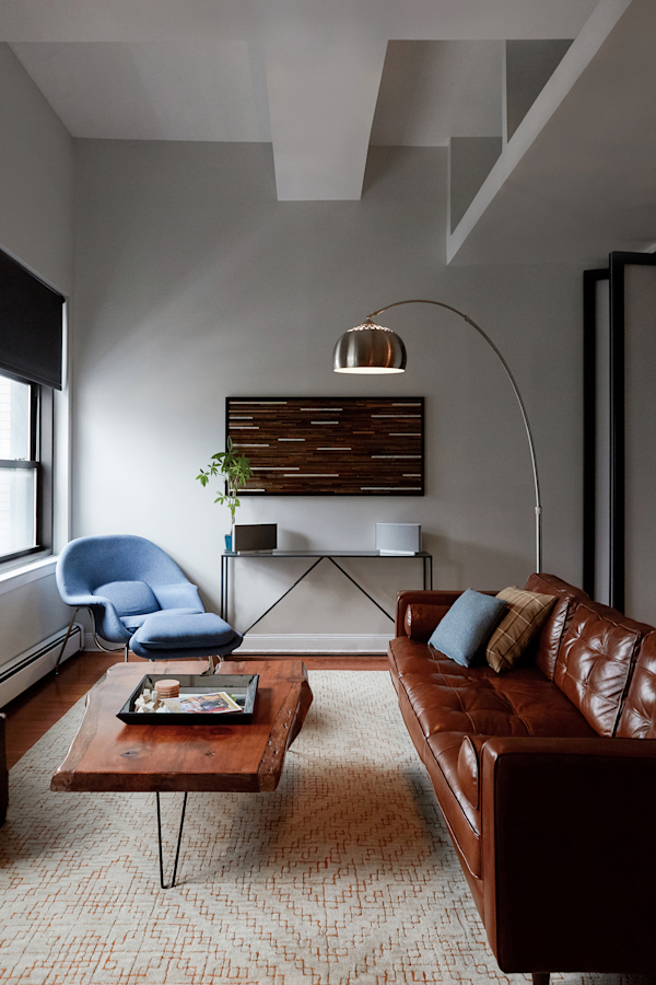 Hell's Kitchen Loft residential interior design by Basicspace. Living room with light blue womb chair, arched floor lamp, leather sofa, soft gray rug with orange and rustic live edge coffee table.