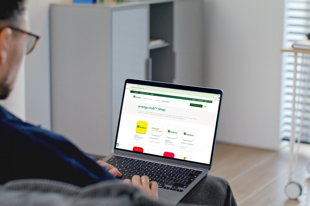 Une personne utilise un ordinateur portable sur un canapé avec l'application AGROLA Energy Club Shop ouverte, qui affiche différentes options d'application.