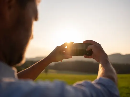 Smartphone ist zentral im Bild, dahinter Sonne erkennbar am Horizont und grüne Landschaft