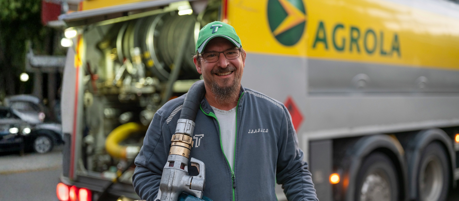 Tankwagenfahrer mit Zapfpistole vor gelbem AGROLA-Tankwagen, trägt grüne Kappe und graue Jacke.