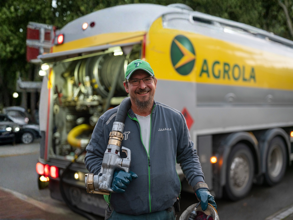 Chauffeur de camion-citerne avec pistolet distributeur devant un camion-citerne jaune AGROLA, portant une casquette verte et une veste grise.